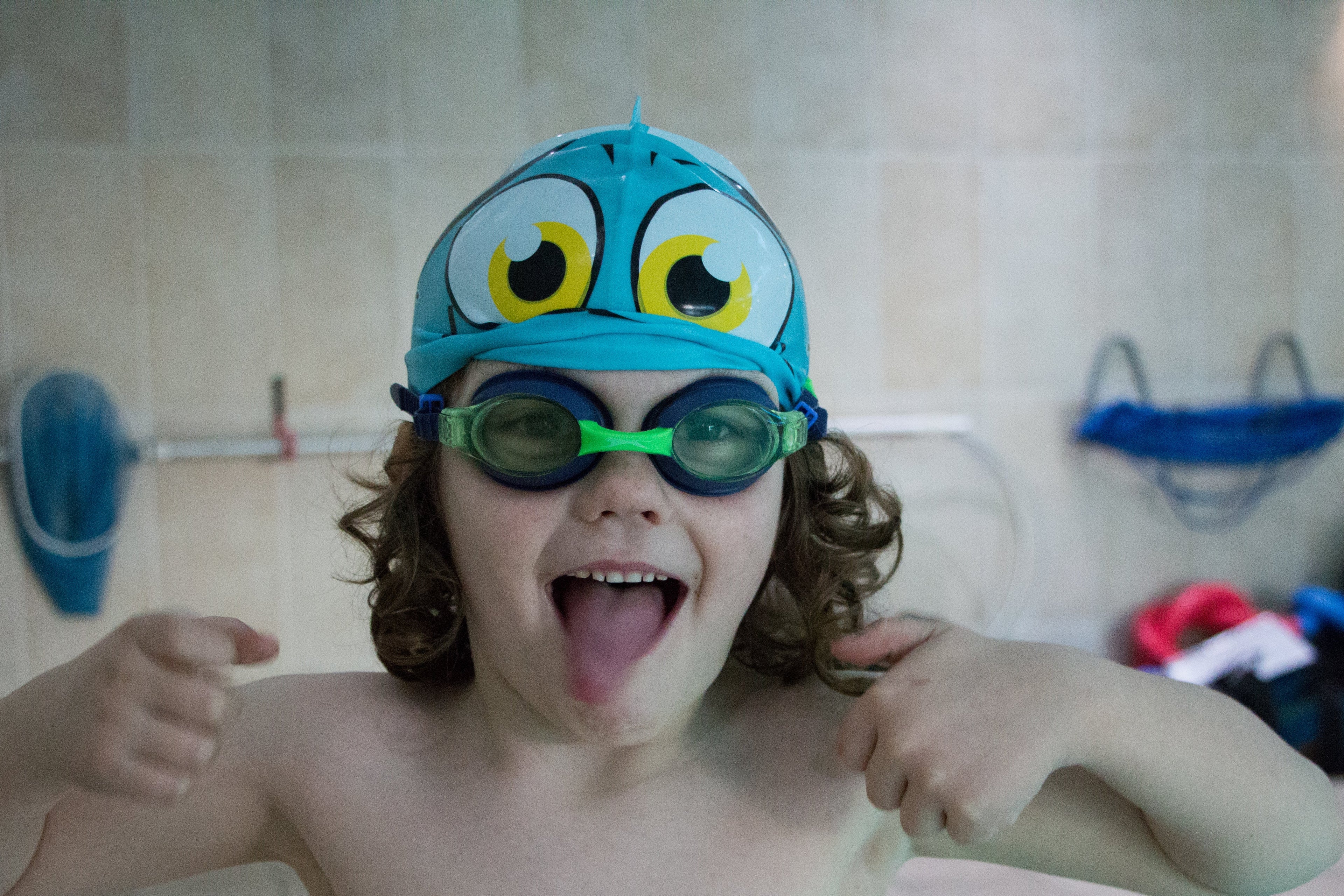 Child wearing swim cap and goggles, with thumbs up, ready for swimming lessons at the poolside.